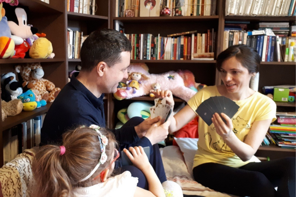 Nicușor Dan, îmbrăcat cu o bluză neagră, soția sa, Mirabela, îmbrăcată cu un tricou galben, în timp ce joacă cărți, având în spate o bibliotecă, iar lângă ei se află fiica lor, care poartă ochelari roz, iar pe cap are o bentiță de flori
