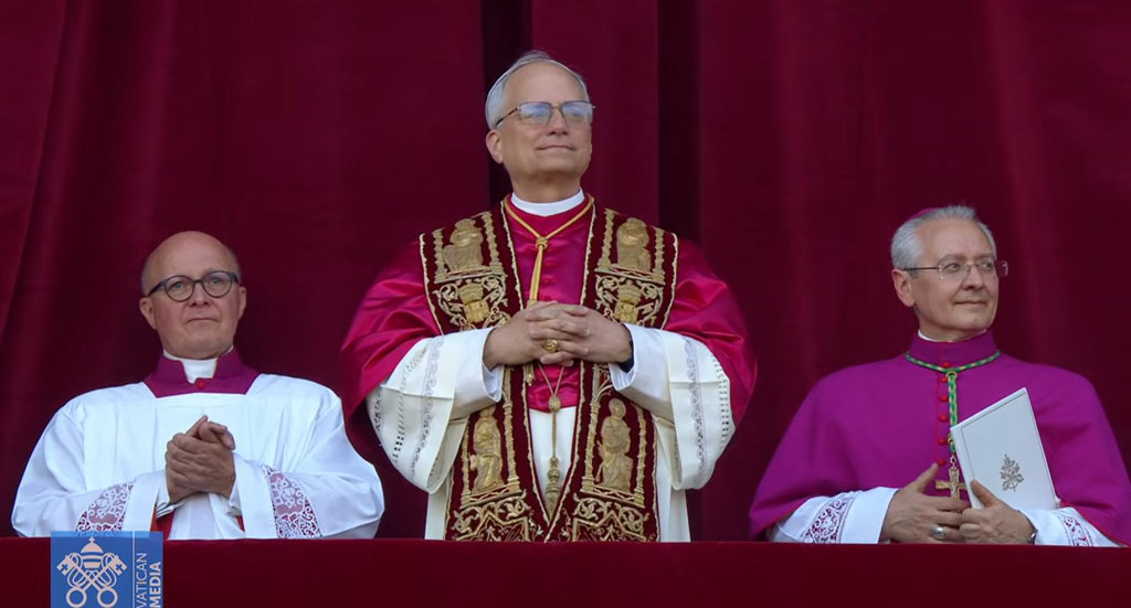 Robert Francis Prevost, noul Papă, iese la balcon îmbrăcat în straiele papale, cu două persoane de-a stânga și de-a dreapta sa.