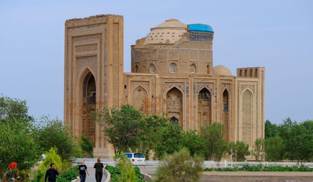Orașul Konye Urgench este din Turkmenistanul de Nord, inclus în Patrimoniul Mondial UNESCO în 2005. In imagine se vede cladirea veche, inconjurată de arbori verzi, oameni care se plimbă