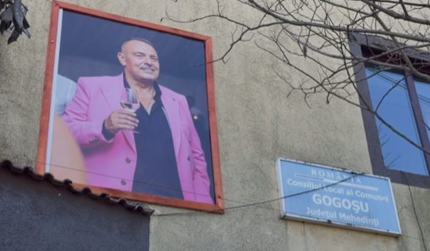 Primarul mort din satul Gogoșu „păzește” primăria cu un pahar de vin în mână, într-un sacou roz: cine a afișat tablourile vesele