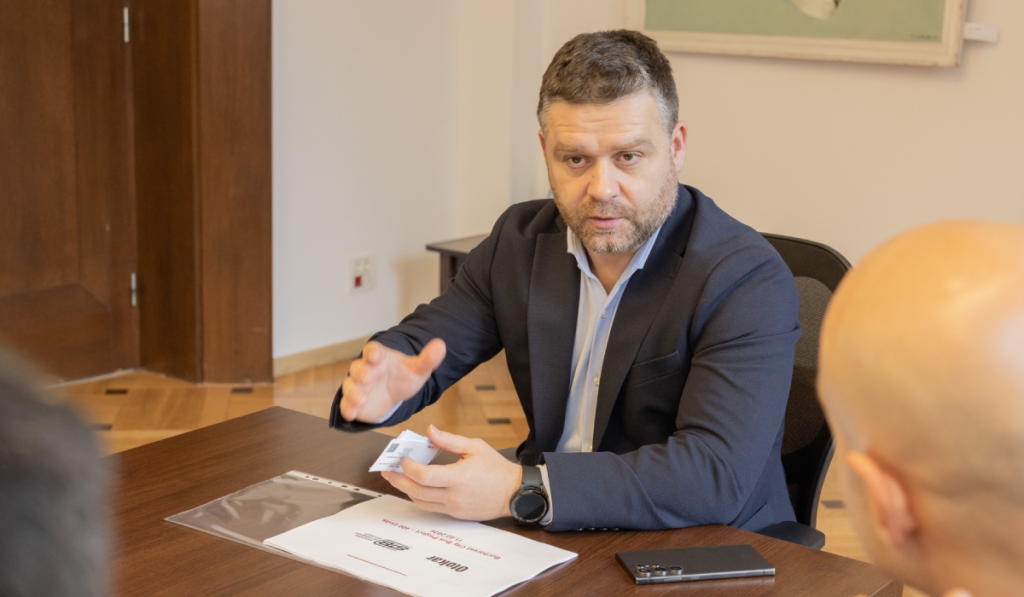 Ciprian Ciucu în costum discută la o masă de birou cu alte persoane, gesticulând, are în față documente și un telefon mobil